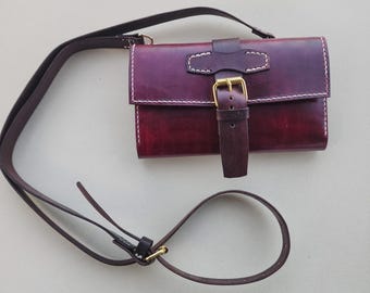 May include: A deep burgundy leather crossbody bag featuring a rectangular flap, white contrast stitching, and a central vertical strap secured with a gold-toned metal buckle. The bag includes a long, adjustable matching leather shoulder strap.
