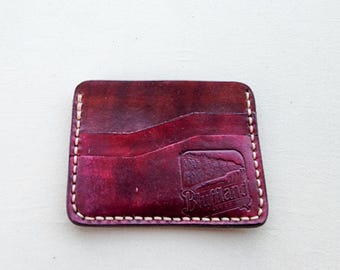 May include: A rectangular burgundy leather card holder featuring a wavy top pocket edge, light-colored contrast stitching along the perimeter, and a debossed logo on the bottom right corner that reads Bluffland.