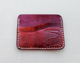 May include: A rectangular dark red leather card holder with rounded corners and light-colored stitching along the edges. The front features a diagonal pocket slot and an embossed logo that reads Bluffland Leather Co.