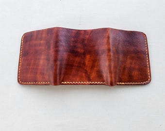 May include: A rectangular brown leather pen case featuring a mottled, textured finish and three distinct vertical molded slots for writing instruments. The edges are secured with visible light-colored stitching along the entire perimeter.