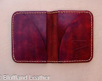 May include: A deep red, hand-stitched leather card holder with rounded corners and two curved pockets. The leather features a mottled, dark-to-light red finish. The word Bluffland is embossed on the bottom right corner of the front panel.