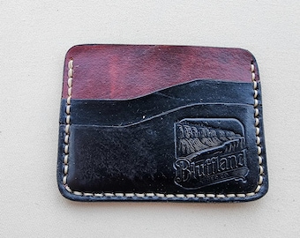 May include: A rectangular leather card holder featuring a two-tone design with a deep red upper section and a black lower section. The piece is stitched with light-colored thread and displays a debossed logo reading Bluffland Leather Co.