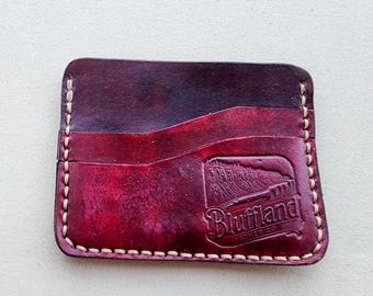 May include: A rectangular leather card holder in a deep burgundy and dark red gradient finish. It features light-colored contrast stitching along the edges and a debossed logo on the bottom right corner that reads Bluffland Leather Co.