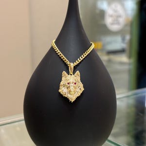May include: A gold wolf pendant with red eyes on a gold chain. The pendant is on a black display stand.