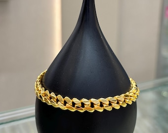 May include: A gold-colored curb chain bracelet displayed on a black teardrop-shaped stand. The bracelet is made of interlocking links and is wrapped around the stand. The background is blurred, suggesting a jewelry store setting.