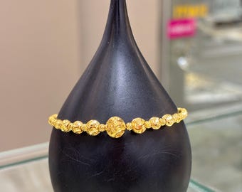 Puede incluir: Un brazalete dorado con una serie de cuentas texturizadas y una cuenta central más grande y elaborada. El brazalete se exhibe sobre un soporte negro en forma de lágrima, mostrando su superficie brillante y reflectante. La joya se encuentra sobre un fondo neutro.