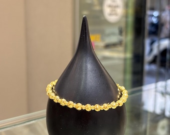 Puede incluir: Un brazalete dorado con cuentas expuesto sobre un soporte negro en forma de lágrima. El brazalete está hecho de pequeñas cuentas redondas y es de un color dorado brillante. El soporte es liso y brillante, contrastando con la textura del brazalete.