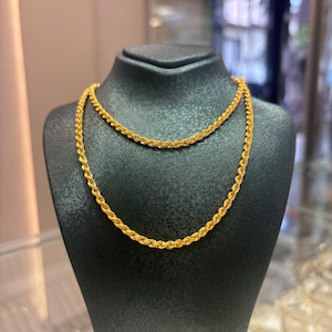 Puede incluir: Un collar de cadena de cuerda dorada exhibido en un maniquí negro. El collar está hecho de eslabones retorcidos, creando una apariencia texturizada. La joyería se presenta en un entorno de tienda, con un fondo borroso.