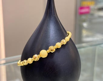 Puede incluir: Un brazalete dorado con una serie de cuentas esféricas texturizadas y lisas. El brazalete se exhibe sobre un soporte negro en forma de lágrima, resaltando su diseño. La joya es un accesorio clásico.