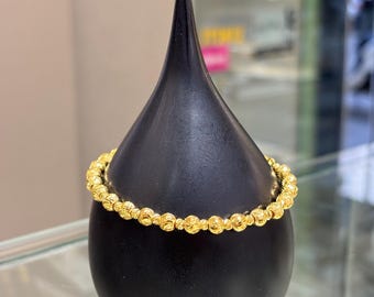 Puede incluir: Un brazalete dorado con cuentas expuesto sobre un soporte negro en forma de lágrima. El brazalete está hecho de pequeñas cuentas redondas con una superficie texturizada. El soporte está sobre una superficie de vidrio, con un fondo borroso.