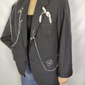 Puede incluir: Una blazer de raya diplom&aacute;tica gris oscuro adornada con cadenas plateadas, imperdibles y una cinta blanca. La blazer presenta un cierre de un solo bot&oacute;n y un bolsillo con una disposici&oacute;n decorativa de alfileres. Una prenda de vestir elegante.