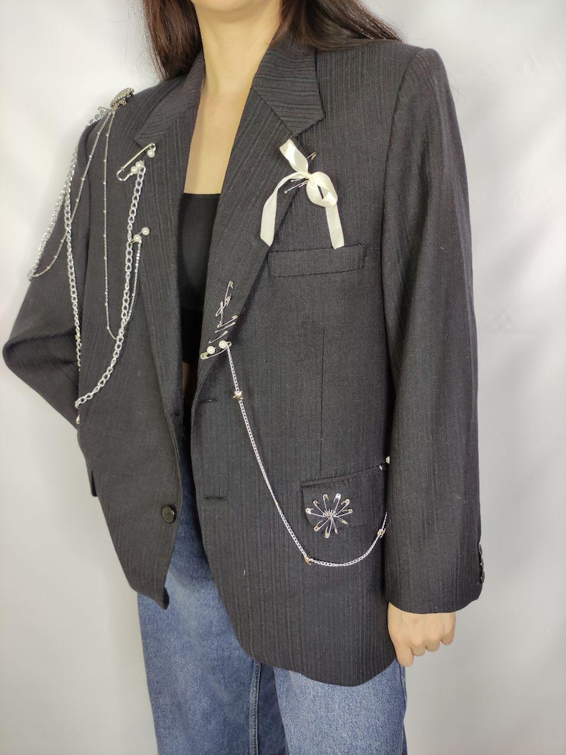 Puede incluir: Una blazer de raya diplom&aacute;tica gris oscuro adornada con cadenas plateadas, imperdibles y una cinta blanca. La blazer presenta un cierre de un solo bot&oacute;n y un bolsillo con una disposici&oacute;n decorativa de alfileres. Una prenda de vestir elegante.