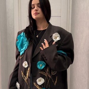 May include: A dark brown blazer adorned with large turquoise and white floral embellishments. Gold embroidered stems and smaller floral accents add detail to the jacket. The blazer is a statement piece.