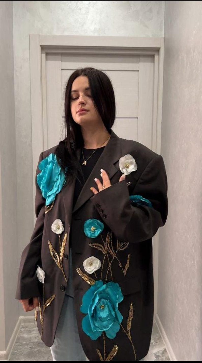 May include: A dark brown blazer adorned with large turquoise and white floral embellishments. Gold embroidered stems and smaller floral accents add detail to the jacket. The blazer is a statement piece.