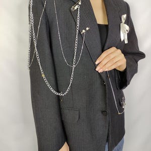 Puede incluir: Un blazer gris oscuro con cadenas plateadas y detalles de perlas. El blazer presenta cadenas decorativas que caen sobre los hombros y el pecho, con un detalle de cinta blanca en la solapa. El blazer tiene un dise&ntilde;o cl&aacute;sico y entallado.
