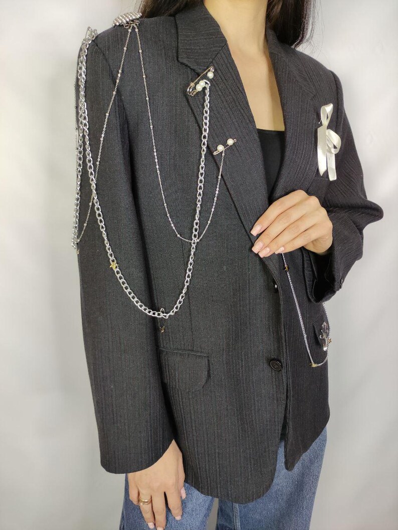 Puede incluir: Un blazer gris oscuro con cadenas plateadas y detalles de perlas. El blazer presenta cadenas decorativas que caen sobre los hombros y el pecho, con un detalle de cinta blanca en la solapa. El blazer tiene un dise&ntilde;o cl&aacute;sico y entallado.