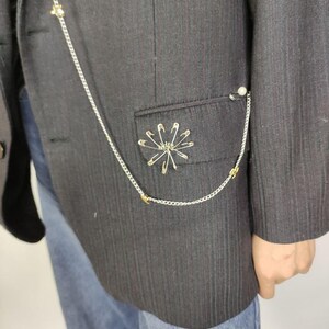 Puede incluir: Una blazer negra con una cadena plateada y adornos de imperdibles. La blazer presenta un dise&ntilde;o decorativo de imperdibles en el bolsillo y una cadena que se conecta a una perla. La blazer se combina con unos vaqueros azules.