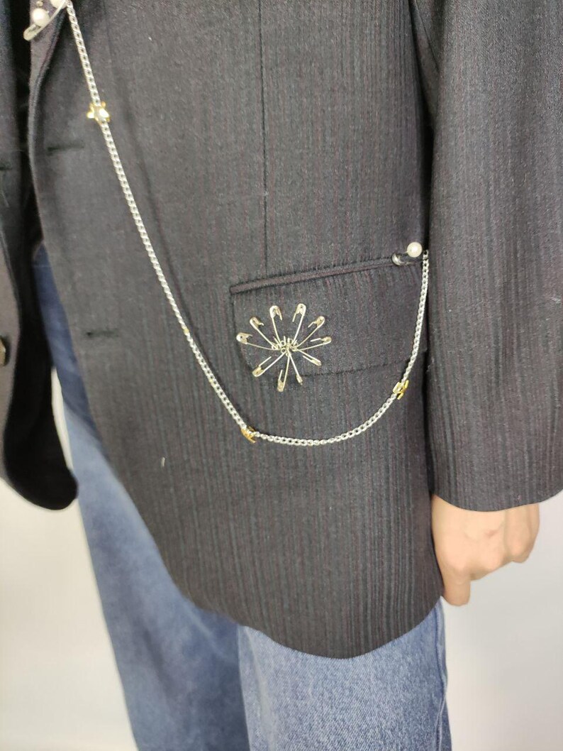 Puede incluir: Una blazer negra con una cadena plateada y adornos de imperdibles. La blazer presenta un dise&ntilde;o decorativo de imperdibles en el bolsillo y una cadena que se conecta a una perla. La blazer se combina con unos vaqueros azules.