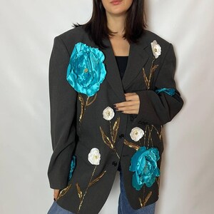 May include: A dark gray blazer adorned with large, turquoise fabric flowers and smaller white floral accents. Gold-colored leaf and stem details add contrast. The blazer has a classic, oversized fit.
