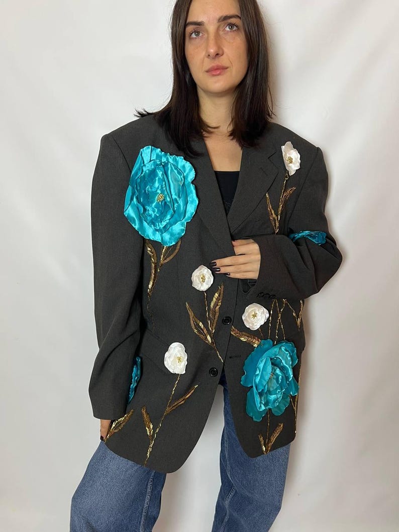 May include: A dark gray blazer adorned with large, turquoise fabric flowers and smaller white floral accents. Gold-colored leaf and stem details add contrast. The blazer has a classic, oversized fit.