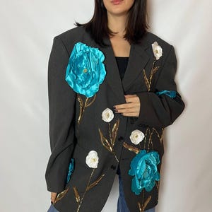 May include: Dark gray blazer adorned with large turquoise and white floral embellishments. The blazer features gold-colored leaf accents and is paired with blue jeans. The jacket has a classic two-button closure.