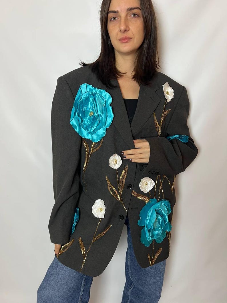 May include: Dark gray blazer adorned with large turquoise and white floral embellishments. The blazer features gold-colored leaf accents and is paired with blue jeans. The jacket has a classic two-button closure.