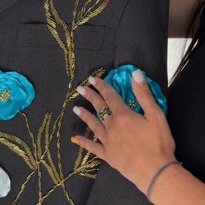 May include: Dark gray blazer with gold beaded floral embroidery and turquoise and white fabric flowers. The blazer has a classic design with a tailored fit. A hand with a ring is touching the blazer.