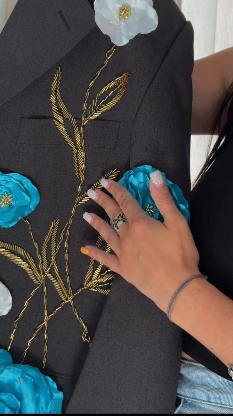 May include: Dark gray blazer with gold beaded floral embroidery and turquoise and white fabric flowers. The blazer has a classic design with a tailored fit. A hand with a ring is touching the blazer.