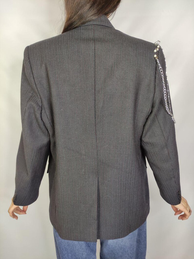 Puede incluir: Un blazer gris oscuro con finas rayas verticales. El blazer presenta una cadena decorativa y detalles de perlas en el hombro. Las mangas tienen pu&ntilde;os abotonados. Un blazer de estilo cl&aacute;sico con un toque moderno.