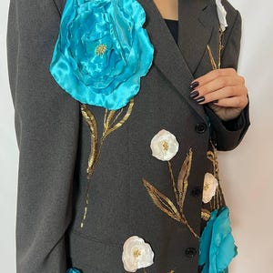 May include: A dark gray blazer adorned with large, turquoise fabric flowers and smaller white flowers. Gold-colored embroidered stems and leaves accent the floral design. The blazer has a classic two-button closure.