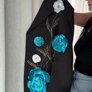 May include: Black blazer adorned with large, vibrant turquoise and white floral embellishments. The jacket features gold embroidered stems and leaves, creating a striking contrast. A stylish, eye-catching piece.