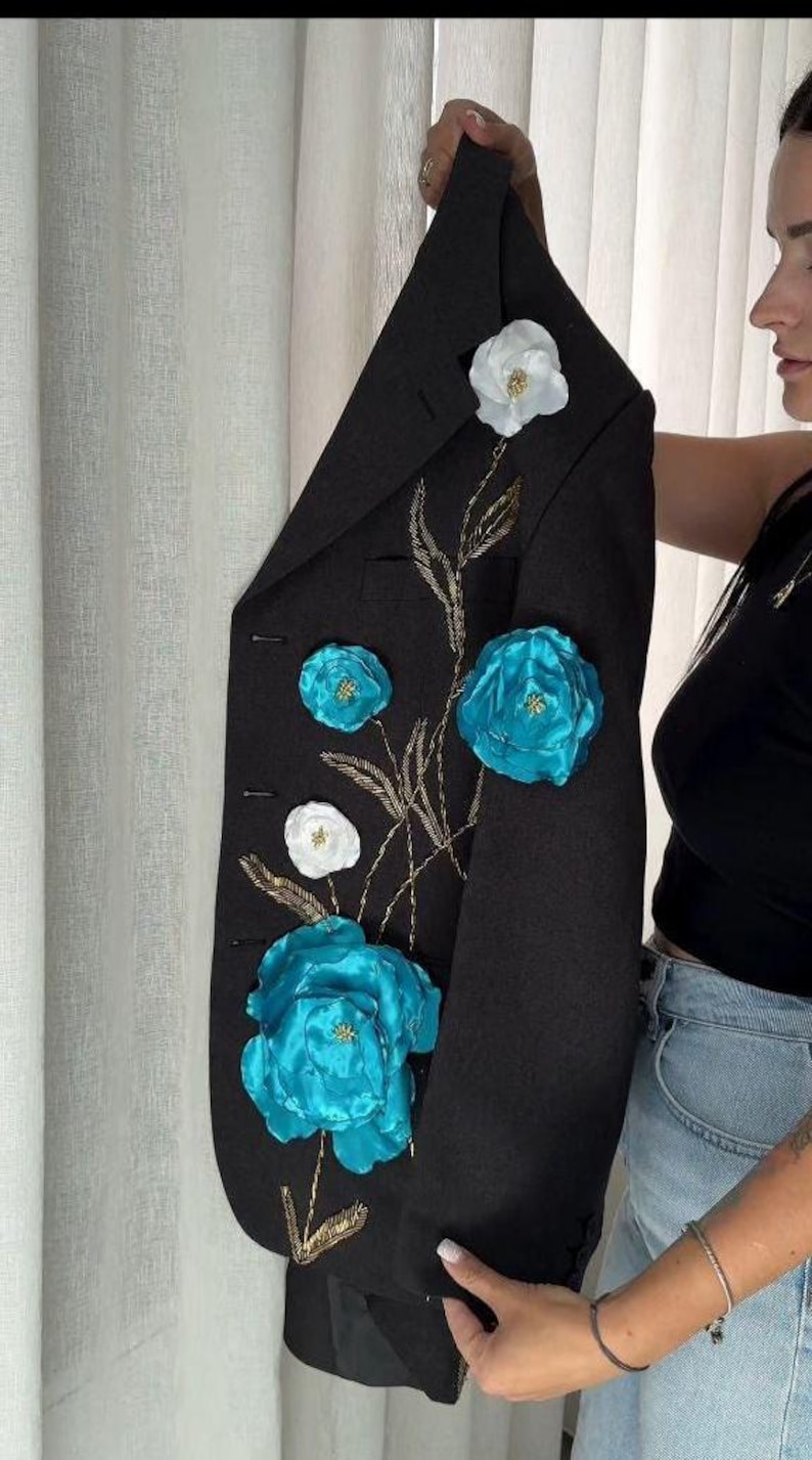 May include: Black blazer adorned with large, vibrant turquoise and white floral embellishments. The jacket features gold embroidered stems and leaves, creating a striking contrast. A stylish, eye-catching piece.
