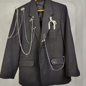 Puede incluir: Un blazer negro con dise&ntilde;o de finas rayas, adornado con cadenas plateadas, alfileres decorativos y una cinta blanca. El blazer tiene dos bolsillos delanteros y est&aacute; colgado de una percha de madera.