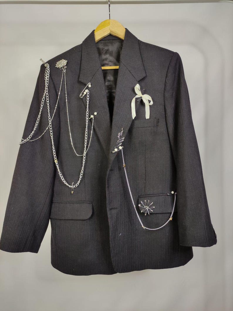 Puede incluir: Un blazer negro con dise&ntilde;o de finas rayas, adornado con cadenas plateadas, alfileres decorativos y una cinta blanca. El blazer tiene dos bolsillos delanteros y est&aacute; colgado de una percha de madera.