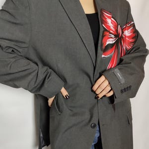 Pode incluir: Um blazer cinza com um grande laço vermelho pintado do lado direito. O blazer tem um fecho de botão único e um bolso do lado esquerdo.