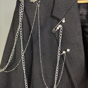 Puede incluir: Un blazer gris oscuro adornado con cadenas plateadas, una broche de diamantes de imitaci&oacute;n y alfileres de seguridad con detalles de perlas. Las cadenas caen por la parte delantera, agregando un toque &uacute;nico y atrevido a la prenda. El blazer est&aacute; colgado en una percha de madera.