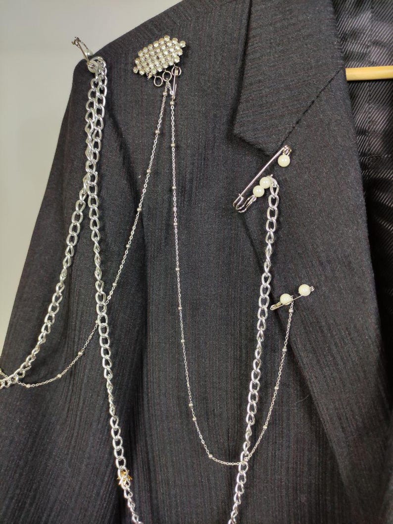 Puede incluir: Un blazer gris oscuro adornado con cadenas plateadas, una broche de diamantes de imitaci&oacute;n y alfileres de seguridad con detalles de perlas. Las cadenas caen por la parte delantera, agregando un toque &uacute;nico y atrevido a la prenda. El blazer est&aacute; colgado en una percha de madera.