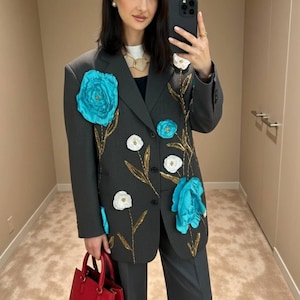 May include: A dark gray blazer and matching pants set adorned with large turquoise and white floral appliqu&eacute;s and gold embroidered stems. The blazer features a classic design with a two-button closure. A red handbag completes the ensemble.
