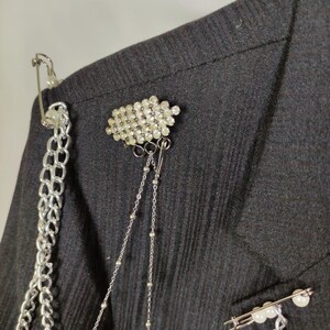 Puede incluir: Primer plano de una blazer negra adornada con broches y cadenas decorativas. La blazer presenta un grupo de pedrer&iacute;a transparente, un imperdible con perlas y cadenas plateadas para un look &uacute;nico y atrevido. La tela tiene una sutil textura acanalada.