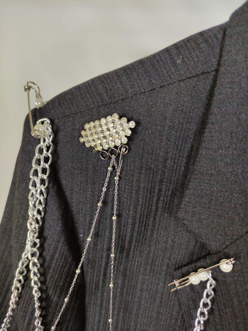 Puede incluir: Primer plano de una blazer negra adornada con broches y cadenas decorativas. La blazer presenta un grupo de pedrer&iacute;a transparente, un imperdible con perlas y cadenas plateadas para un look &uacute;nico y atrevido. La tela tiene una sutil textura acanalada.