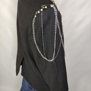 Puede incluir: Blazer negro con cadenas plateadas y adornos de hombro de perlas. La chaqueta presenta una tela texturizada y un dise&ntilde;o cl&aacute;sico a medida. Cadenas decorativas y imperdibles a&ntilde;aden un estilo &uacute;nico y atrevido a la prenda.