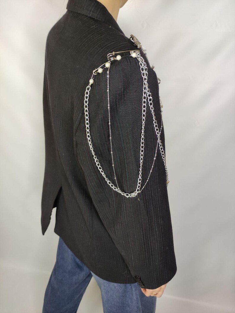 Puede incluir: Blazer negro con cadenas plateadas y adornos de hombro de perlas. La chaqueta presenta una tela texturizada y un dise&ntilde;o cl&aacute;sico a medida. Cadenas decorativas y imperdibles a&ntilde;aden un estilo &uacute;nico y atrevido a la prenda.