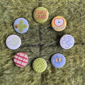 Op de afbeelding: Een set van acht ronde buttons met diverse ontwerpen, waaronder een groene button met de tekst TOUCH GRASS, een gele button met een appel en de tekst FRESH, een paarse button met een bloem en een rood-wit geblokte button.