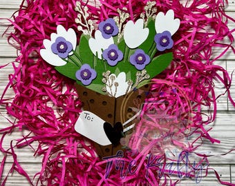 May include: A paper craft bouquet of white flowers with purple centers, arranged in a brown paper cone with a white tag that says "To:". The bouquet is surrounded by pink crinkle paper.
