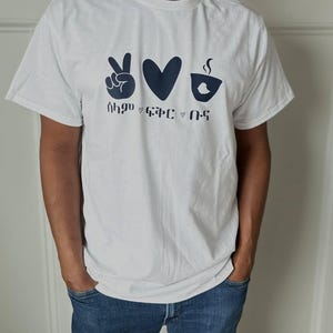 May include: A white t-shirt with a navy blue graphic design. The design includes a peace sign, a heart, and a coffee cup with steam, along with text below the symbols. The shirt is short-sleeved and worn by a person.