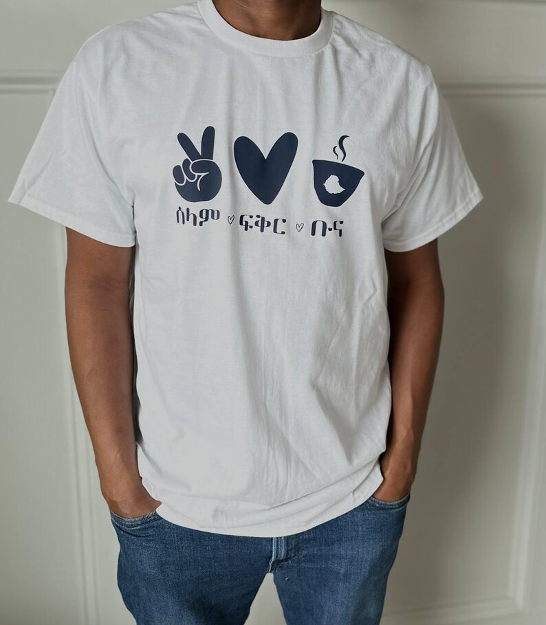 May include: A white t-shirt with a navy blue graphic design. The design includes a peace sign, a heart, and a coffee cup with steam, along with text below the symbols. The shirt is short-sleeved and worn by a person.