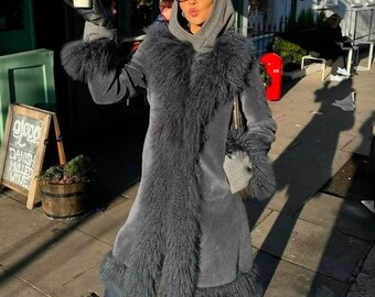May include: A charcoal gray long coat featuring a thick, shaggy faux fur trim along the collar, cuffs, and hem. The coat is worn over a gray hooded sweatshirt and dark blue denim jeans. The outfit includes black leather gloves and oval-shaped glasses.