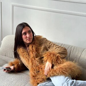 May include: A brown suede jacket with long sleeves and a fluffy, light brown fur trim around the collar and cuffs. The jacket is worn over blue jeans. The jacket has a V-neck design.