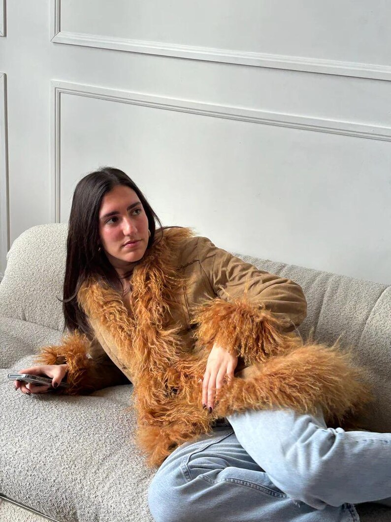 May include: A brown suede jacket with long sleeves and a fluffy, light brown fur trim around the collar and cuffs. The jacket is worn over blue jeans. The jacket has a V-neck design.