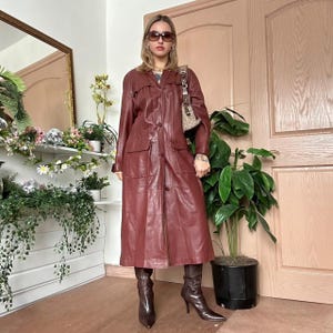 May include: A burgundy leather trench coat with a button-up front, pockets, and a longline silhouette. The coat is paired with matching knee-high boots and a patterned handbag. The outfit is worn by a person with sunglasses.